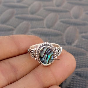 May include: A silver ring featuring an oval-shaped iridescent abalone shell inlay. The setting is framed by a twisted rope-style border with decorative spiral accents on each side. The text "BoheRingArt" is visible on the surface of the shell.