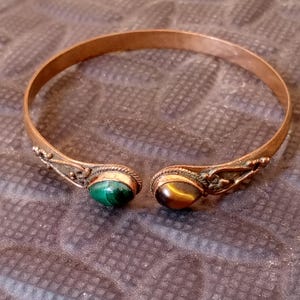 May include: Copper-toned cuff bracelet with decorative ends. One end features a green malachite stone, the other a brown tiger's eye stone. The bracelet has an open design and is likely for a wrist size of approximately 6-8 inches.