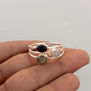 Op de afbeelding: Een set van drie zilveren ringen, elk met een ovale edelsteen. De edelstenen zijn zwart, iriserend en groen. De ringen zijn op elkaar gestapeld, waardoor een gelaagd effect ontstaat.