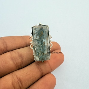 May include: A rectangular ring with a silver band and a large, rectangular stone. The stone has a mottled pattern of gray, green, and white. The ring is on a hand. The text "BohoRingArt" is visible.