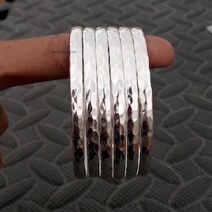 Puede incluir: Un conjunto de seis brazaletes de color plateado con una superficie martillada y texturizada. Cada brazalete tiene un acabado metálico reflectante con hendiduras en el exterior. El texto BohoRingArt es visible en el primer brazalete.