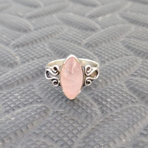 Könnte beinhalten: Silberring mit einem großen, ovalen Rosenquarz-Edelstein. Der Ring hat verzierte, wirbelnde Designs an den Seiten des Bandes. Der Rosenquarz hat eine zarte rosa Farbe und ist in einem Silberband gefasst.