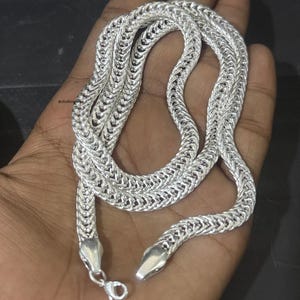 May include: A silver chain necklace with a woven design. The necklace is displayed on a hand, highlighting its intricate pattern and the shiny metal. The chain has a lobster clasp closure.