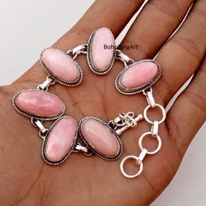 May include: A silver bracelet with oval, pale pink gemstones. Each gemstone is set in a silver bezel with a beaded edge. The bracelet is connected by silver links and has a clasp closure. The bracelet is displayed on a hand.