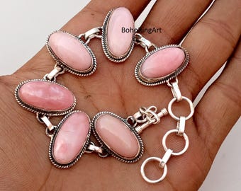 May include: A silver bracelet with oval, pale pink gemstones. Each gemstone is set in a silver bezel with a beaded edge. The bracelet is connected by silver links and has a clasp closure. The bracelet is displayed on a hand.