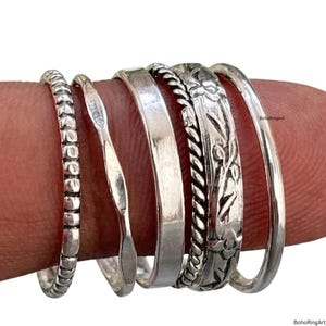 Puede incluir: Una colección de seis anillos de plata de varios diseños. Los anillos incluyen una banda con cuentas, una banda martillada, una banda lisa, una banda de cuerda trenzada, una banda grabada floral y una banda simple. Los anillos se muestran en un dedo.