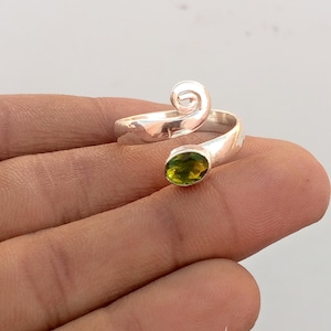 Könnte beinhalten: Ein silberfarbener Ring mit einem Wirbeldesign und einem grünen, ovalen Edelstein. Der Ring umschließt den Finger mit einem offenen Design und einer polierten Oberfläche. Der Edelstein hat eine leuchtend grüne Farbe.