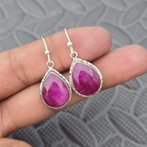 May include: A pair of silver teardrop earrings with large, vibrant magenta gemstones. The gemstones are set in a silver bezel with a textured border. The earrings have a simple hook closure.