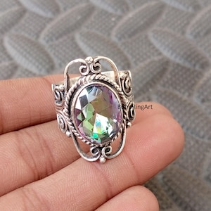 Puede incluir: Un anillo de plata con una piedra preciosa ovalada e iridiscente. El anillo tiene un diseño de filigrana ornamentado alrededor de la piedra y en la banda. La piedra preciosa muestra una gama de colores, incluyendo verde, morado y amarillo. El anillo es una joya.