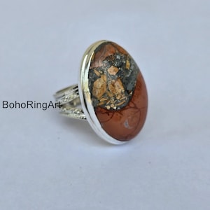 May include: A silver ring with an oval-shaped stone. The stone is a mix of brown, black, and tan colors. The ring band has a decorative design. The text "BohoRingArt" is visible on the left side.