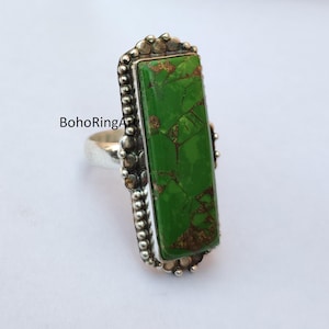 May include: A silver ring featuring a large, rectangular green stone with brown veining. The ring has a detailed setting with small, round embellishments. The text "BohoRingArt" is visible on the ring.