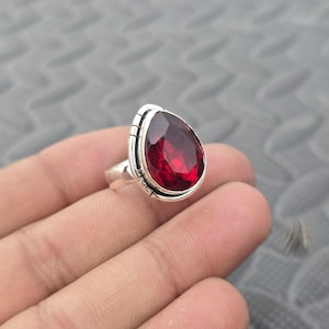 May include: A silver ring with a large, teardrop-shaped, faceted red gemstone. The gemstone is the focal point, set in a detailed silver band with vertical lines. The ring is displayed on a hand, showcasing its size and design.