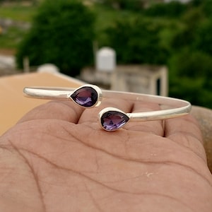 Peut inclure: Un bracelet ouvert en argent doté de deux pierres précieuses violettes à facettes en forme de poire à chaque extrémité. La bande métallique présente une finition lisse et polie avec un petit poinçon 925 sur la face interne.