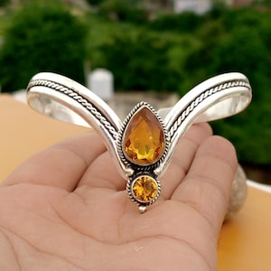 May include: A silver bracelet in a tiara design, adorned with two amber gemstones. The larger, pear-shaped stone is centrally placed, with a smaller, round stone beneath. The bracelet has a textured, rope-like pattern along the edges.