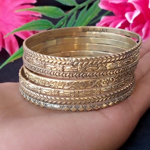 May include: A stack of five gold-toned metal bangles featuring various embossed patterns, including floral motifs, geometric shapes, and braided textures. The bracelets are arranged in a vertical column, showcasing intricate detailing on each band.