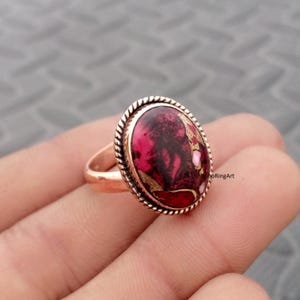 Peut inclure: Une bague en cuivre avec une pierre ovale au motif marbré magenta foncé et noir, rehaussée d'inclusions dorées métalliques. La pierre est sertie dans un bord en métal torsadé en forme de corde, et l'anneau présente une finition lisse et polie.