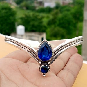 May include: A silver-toned V-shaped cuff bracelet featuring a central pear-shaped blue gemstone and a smaller round blue gemstone below it. The metal band has a twisted rope texture along the edges and a polished finish on the inner surface.