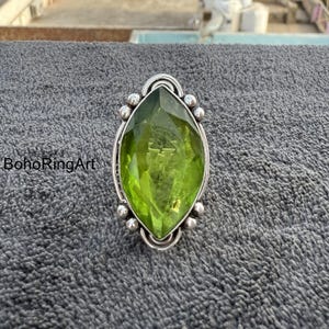 Könnte beinhalten: Ein silberner Ring mit einem großen, ovalförmigen, hellgrünen Edelstein. Der Edelstein ist facettiert und von einer silbernen Fassung mit kleinen, runden Akzenten umgeben. Der Text "BohoRingArt" ist sichtbar.