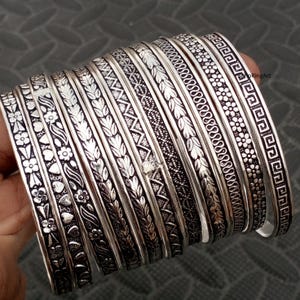 Pode incluir: Um conjunto de dez pulseiras de metal prateado com vários padrões em relevo, incluindo motivos florais, corações, folhas, ziguezagues geométricos, texturas de renda intrincadas e uma borda grega, com um acabamento oxidado escuro.