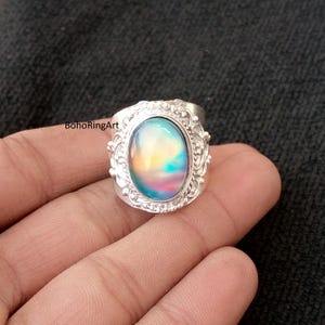 May include: A silver ring with an oval, iridescent gemstone. The gemstone displays a blend of colors, including blue, yellow, and pink. The ring has an ornate, filigree design around the gemstone and the text "BohoRingArt".