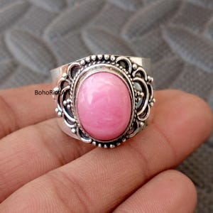 May include: A silver ring featuring a light pink oval gemstone. The ring has an elaborate, detailed setting with a decorative border. The gemstone is the central element, set within a bezel. This ring is likely a piece of jewellery.
