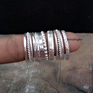 Può includere: Una collezione di anelli d'argento con vari design, tra cui corde intrecciate, motivi incisi e fasce lisce. Gli anelli sono esposti su un dito su sfondo scuro. Gli anelli sono un gioiello.