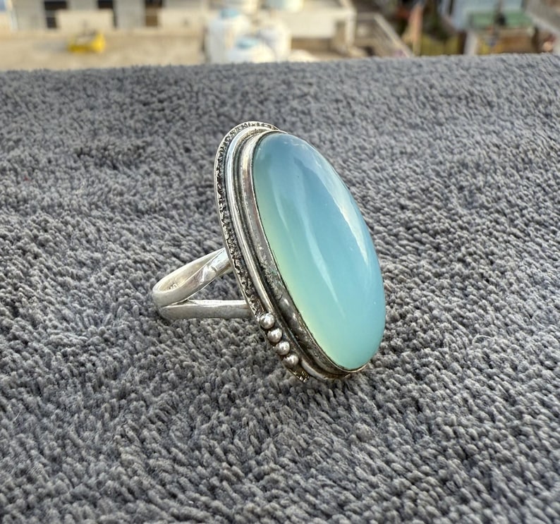 K&ouml;nnte beinhalten: Ein Silberring mit einem gro&szlig;en, ovalen, hellblauen Edelstein. Der Edelstein ist in einer detaillierten Silberfassung mit einem strukturierten Rand gefasst. Der Ring hat ein schlichtes Band, das den Stein hervorhebt.