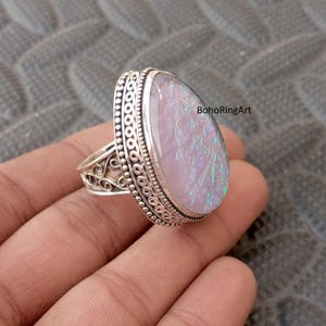 Op de afbeelding: Een grote, ovale ring met een iriserende steen in roze, wit en groen. De ring is gezet in zilver metaal met decoratieve details rond de steen en op de band. De tekst "BohoRingArt" is zichtbaar. Een opvallend sieraad.