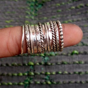 May include: A collection of ten silver-toned metal rings arranged in a row. The rings feature various textures and patterns, including smooth bands, twisted rope designs, etched floral motifs, and beaded edges, all displayed together on a finger.
