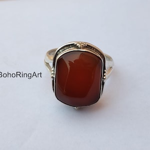 Può includere: Un anello d'argento con una grande pietra rettangolare marrone-rossastra. L'anello ha un'incastonatura decorativa attorno alla pietra e una fascia semplice. Il testo "BohoRingArt" è visibile sul lato sinistro dell'immagine.