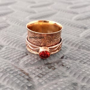 Peut inclure: Une bague rotative de couleur dorée avec une pierre rouge. La bague présente des gravures florales sur le bracelet et le texte "BohoRingArt". La pierre précieuse est d'un rouge profond et est sertie dans un sertissage à lunette.