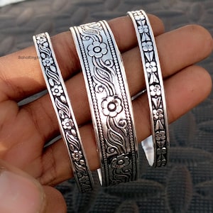 Puede incluir: Tres brazaletes de metal plateado con intrincados patrones florales y de enredaderas en relieve con detalles en negro oxidado. Los brazaletes varían en ancho, siendo la pieza central la más ancha, todos con un motivo botánico consistente.