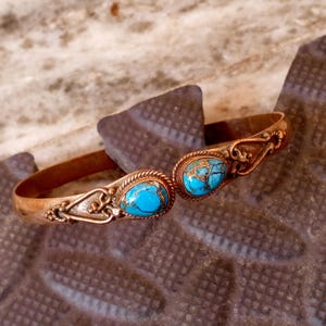 Peut inclure: Un bracelet manchette de couleur cuivre avec deux pierres ovales turquoise. Les pierres présentent un motif bleu et or. Le bracelet comporte des détails de filigrane décoratifs de chaque côté des pierres et le long du bracelet.