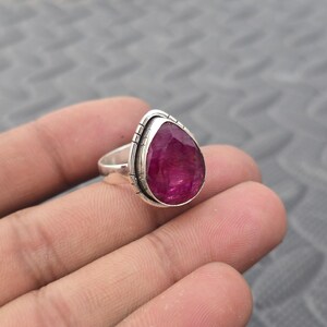 Könnte beinhalten: Ein Silberring mit einem großen, birnenförmigen, tiefroten Edelstein. Der Edelstein ist in einer Lünettenfassung mit dekorativem Rand gefasst. Der Ring hat ein schlichtes, poliertes Band, das die Farbe des Steins hervorhebt.