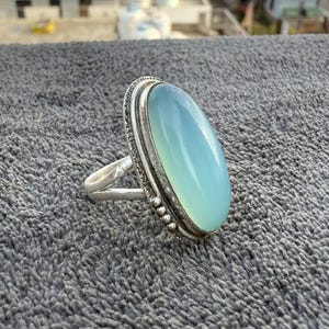 K&ouml;nnte beinhalten: Ein Silberring mit einem gro&szlig;en, ovalen, hellblauen Edelstein. Der Edelstein ist in einer detaillierten Silberfassung mit einem strukturierten Rand gefasst. Der Ring hat ein schlichtes Band, das den Stein hervorhebt.