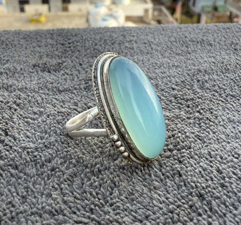 K&ouml;nnte beinhalten: Ein Silberring mit einem gro&szlig;en, ovalen, hellblauen Edelstein. Der Edelstein ist in einer detaillierten Silberfassung mit einem strukturierten Rand gefasst. Der Ring hat ein schlichtes Band, das den Stein hervorhebt.