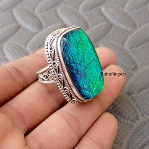Puede incluir: Un anillo de plata con una piedra grande, rectangular, verde y azul iridiscente. La piedra tiene una apariencia texturizada. El anillo tiene un diseño ornamental de filigrana alrededor de la piedra y en la banda. El texto "BohoRingArt" es visible.