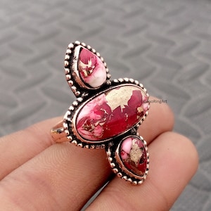 Peut inclure: Une bague en cuivre ornée de trois pierres au motif marbré rouge foncé, rose et blanc, rehaussées d'inclusions dorées métalliques. La pierre centrale ovale est entourée de deux pierres en forme de goutte, serties dans du métal perlé.