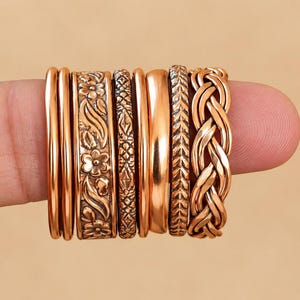 May include: A set of seven copper-toned rings featuring various designs, including two plain bands, one with a floral pattern, one with a geometric diamond motif, one smooth band, one with a chevron texture, and one with a braided design.