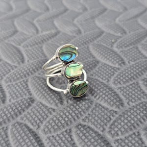 Puede incluir: Un anillo de plata con tres piedras ovaladas de concha de abulón. Las piedras muestran iridiscencias azules, verdes y marrones. El anillo tiene un diseño multibanda con pequeñas cuentas de plata.