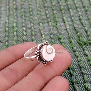 May include: A silver ring with a white and tan spiral design. The ring has a decorative setting around the central stone and is set against a green background. The ring is a piece of jewelry.
