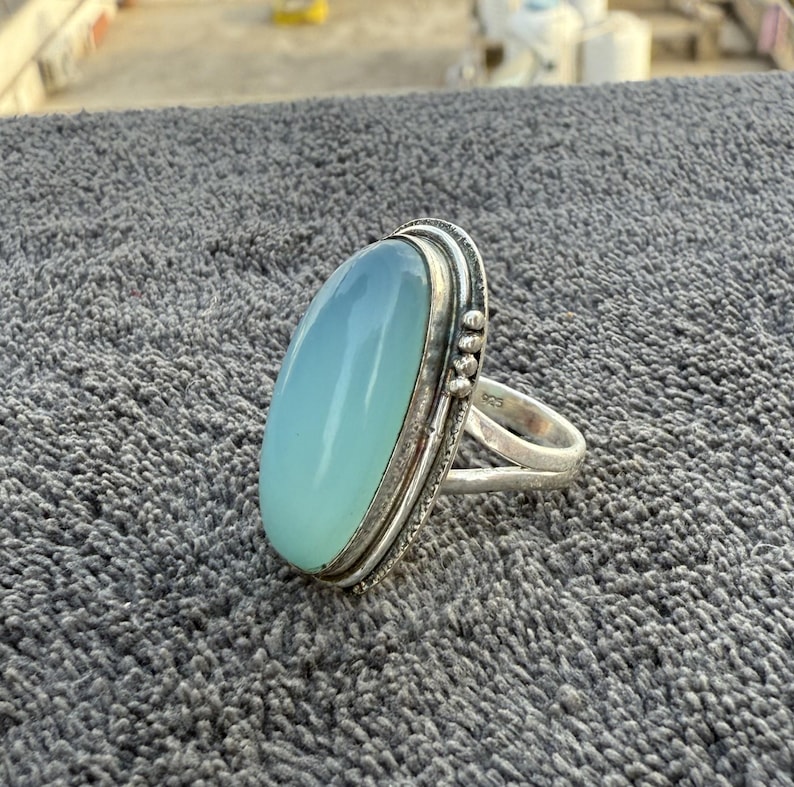 K&ouml;nnte beinhalten: Ein Silberring mit einem gro&szlig;en, ovalen, hellblauen Edelstein. Der Edelstein hat eine glatte, polierte Oberfl&auml;che und ist in einem detaillierten Silberband gefasst. Der Ring wird auf einer strukturierten grauen Oberfl&auml;che pr&auml;sentiert.