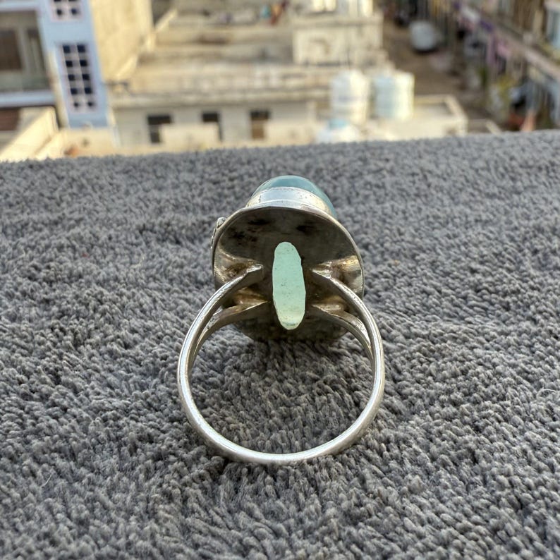K&ouml;nnte beinhalten: Ein silberner Ring mit einem gro&szlig;en, ovalen, hellblauen Edelstein. Der Ring hat eine detaillierte Fassung und ein geteiltes Banddesign. Der Edelstein ist der Mittelpunkt, mit einem subtilen, durchscheinenden Aussehen. Der Ring ist vor einem strukturierten grauen Hintergrund platziert.
