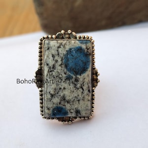 May include: A rectangular ring with a k2 stone displaying blue and grey patterns. The ring has a gold-coloured, ornate setting with a beaded border and decorative accents. The text "BohoRingArt" is visible on the side of the ring.