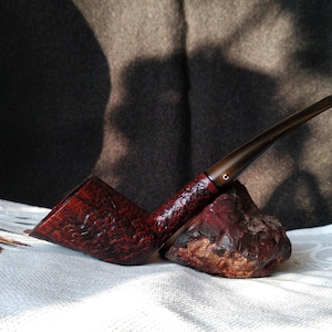 Pode incluir: Um cachimbo de madeira marrom escuro com acabamento texturizado. O cachimbo está apoiado em uma superfície branca com uma rocha vermelha.