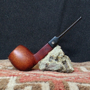 Pode incluir: Um cachimbo de madeira marrom com um caule preto apoiado em uma pedra. O cachimbo está em um tapete estampado.