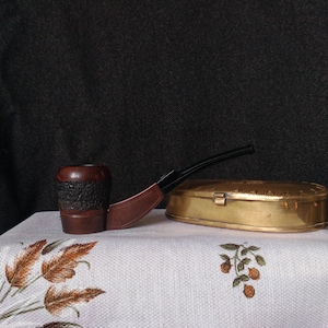 May include: A wooden pipe with a brown bowl and a black stem, resting on a white tablecloth with a gold metal box next to it.