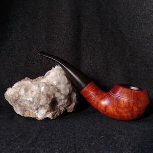 Könnte beinhalten: Eine Holzpfeife mit einem dunkelbraunen Mundstück und einem rotbraunen Kopf. Die Pfeife liegt auf einem rauen, cremefarbenen Stein vor dunklem Hintergrund. Der Kopf hat ein geschnitztes Design.