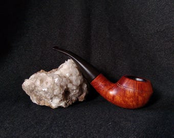 May include: A wooden tobacco pipe with a dark brown stem and a reddish-brown bowl. The pipe rests on a rough, off-white rock against a dark background. The bowl has a carved design.