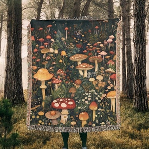 May include: A woven tapestry throw blanket featuring a dark green background with a colorful illustration of mushrooms and flowers. The blanket has a fringe border.